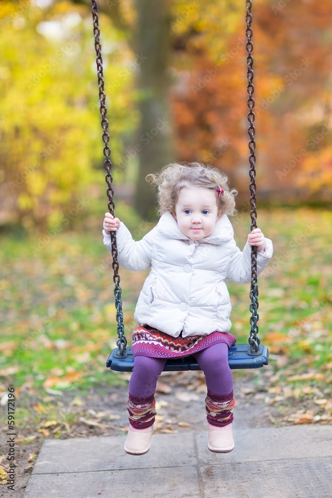 Cute toddler girl playing on a swing with beautiful autumn trees