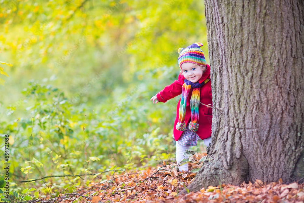 Sweet funny baby girl in a red coat and colorful hat in a park