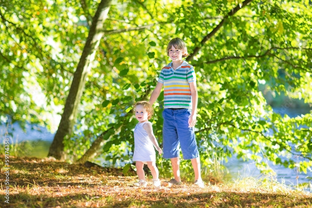 Fototapeta premium Cute school boy holding his baby sister playing at a river shore