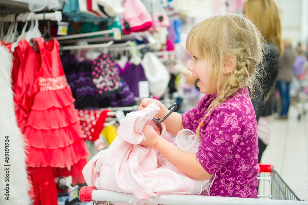 Adorable girl on shoppping cart select pink dress in supermarket