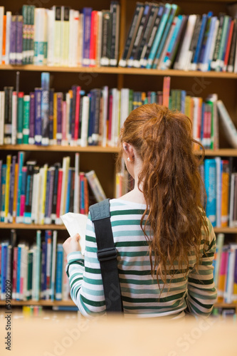 Redhead student reading book from shelf standing in library