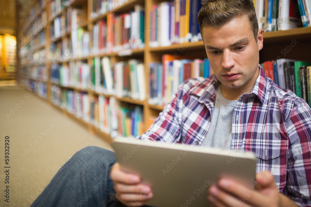 Serious young student sitting on library floor using tablet Serious young student sitting on library floor using tablet