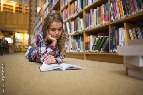 Pretty focused student lying on library floor reading book