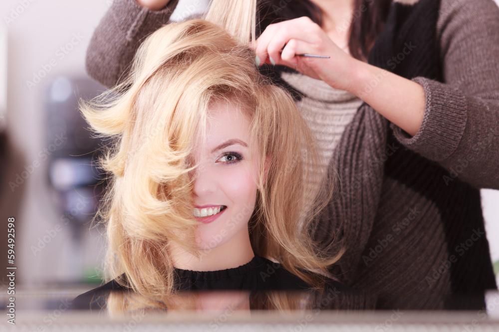 Fototapeta premium Smiling girl with blond wavy hair by hairdresser in beauty salon