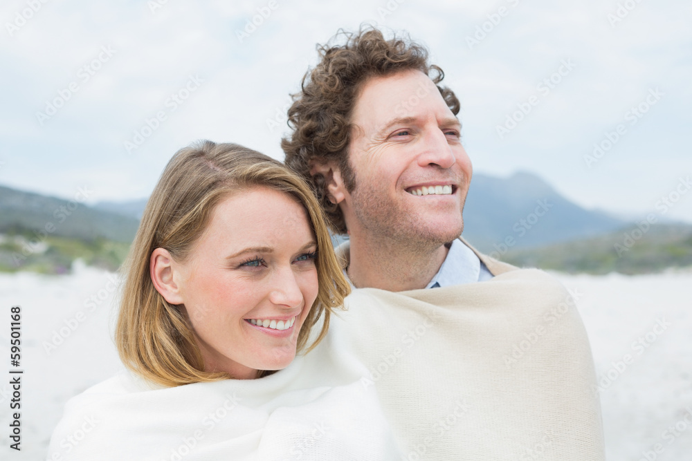 Smiling couple wrapped in blanket at beach - obrazy, fototapety, plakaty 