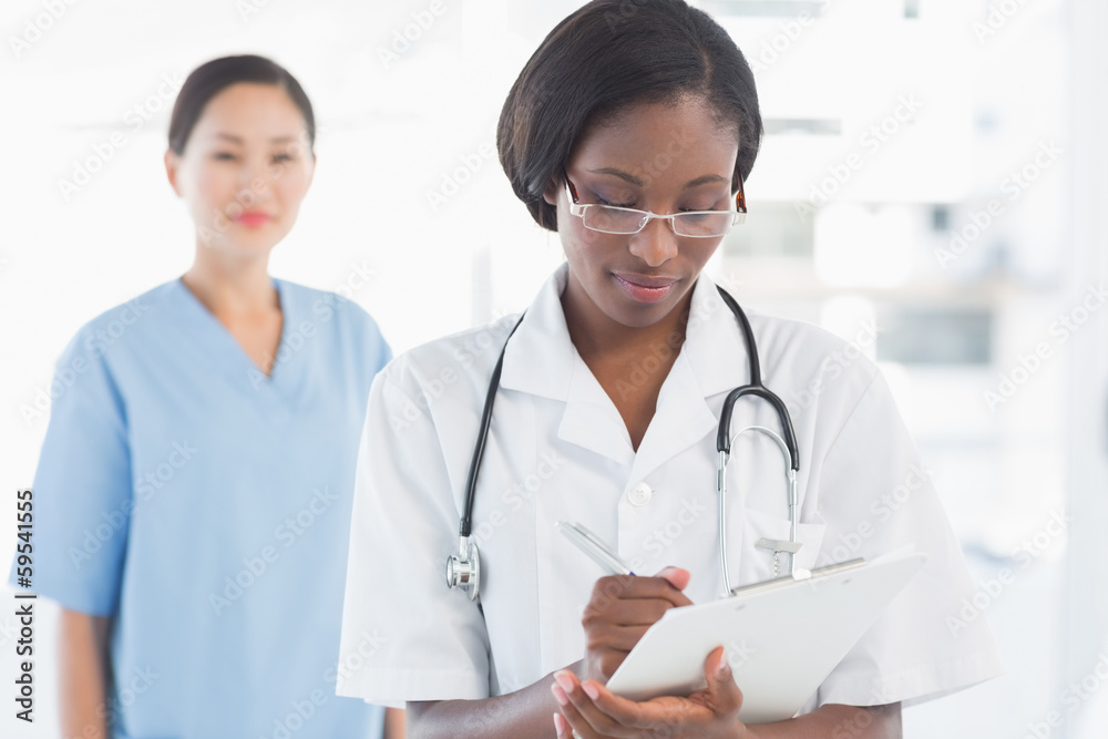 Female doctor writing report with surgeon in background Female doctor writing report with surgeon in background