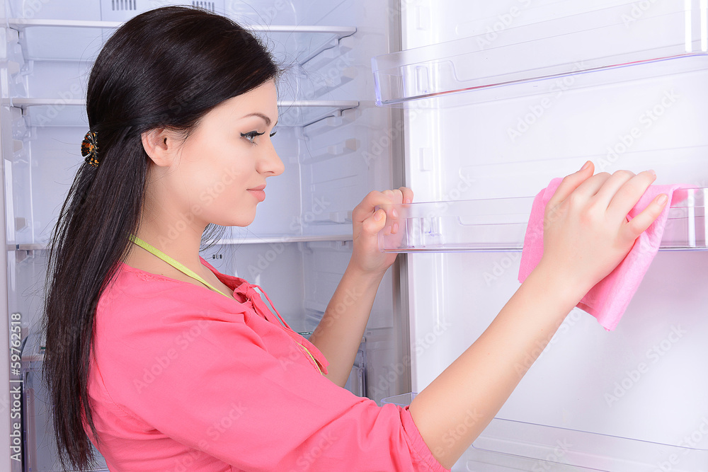 Beautiful young woman washing fridge in kitchen