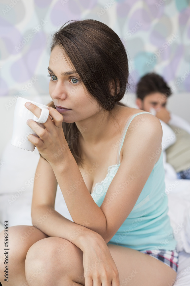 Young woman having coffee on bed with man lying in background Young woman having coffee on bed with man lying in background