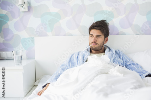 Sick man with thermometer in mouth reclining on bed at home