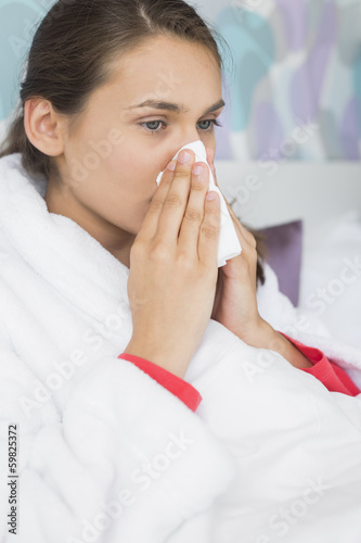 Young woman suffering from cold blowing nose in bedroom