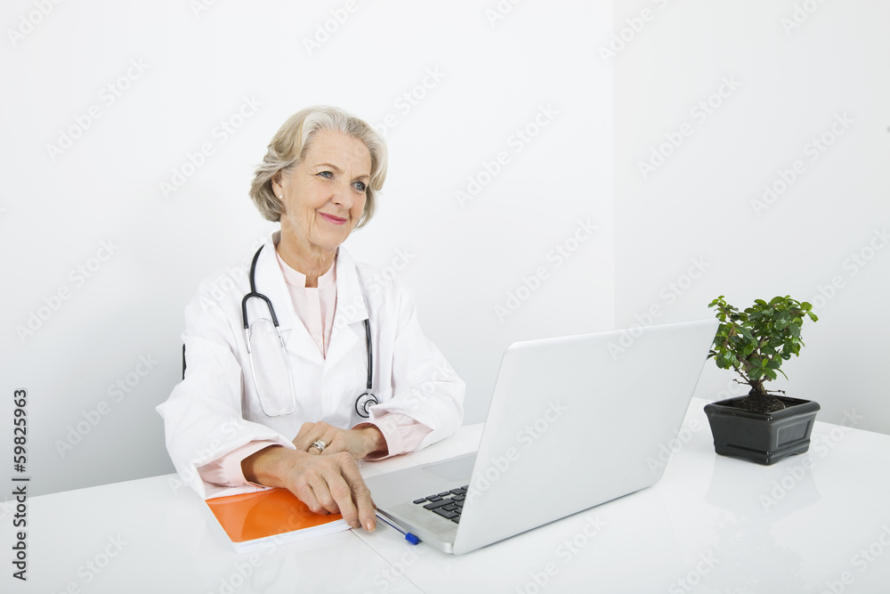 Thoughtful senior female doctor with laptop at desk in clinic Thoughtful senior female doctor with laptop at desk in clinic