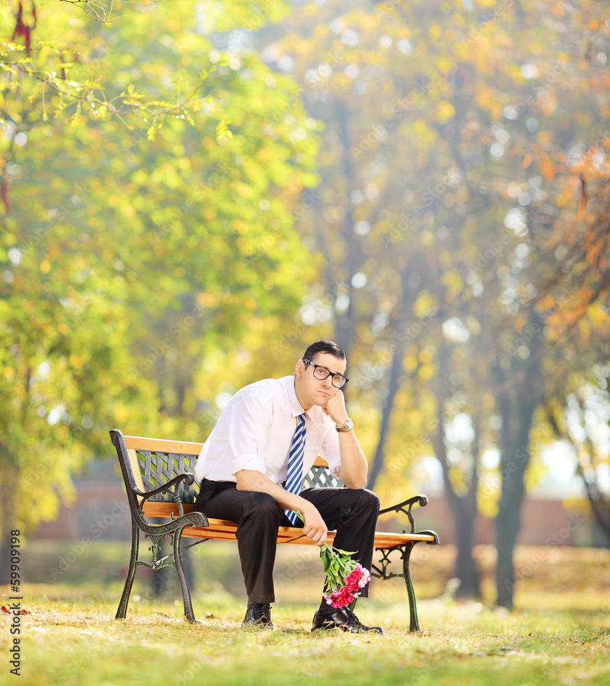 Sad young man holding flowers and sitting on a bench in a park Sad young man holding flowers and sitting on a bench in a park