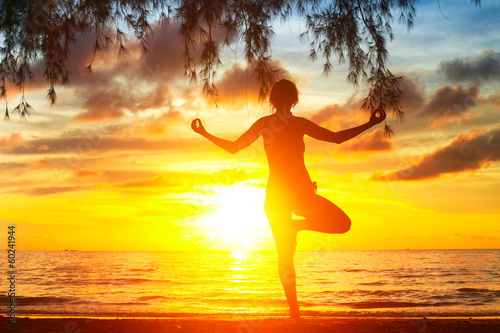 Young woman silhouette practicing yoga on the beach at sunset.