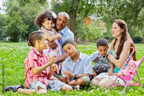 Mixed race family of six rests sitting on grass in summer park