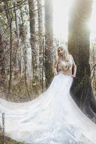 Blonde bride angel in long white skirt standing in sunny forest