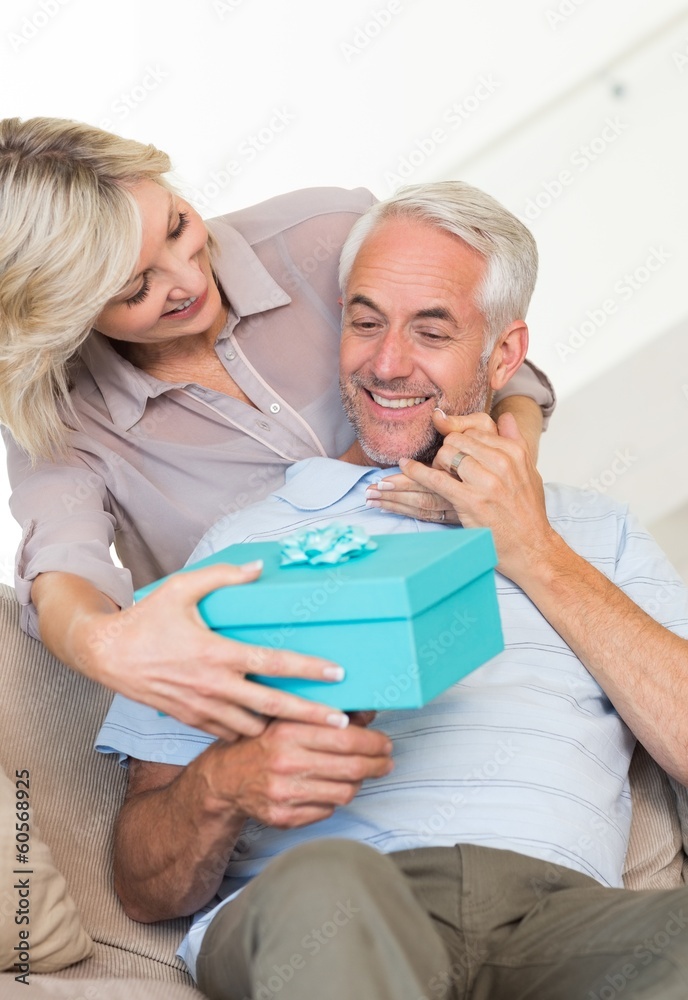 Obraz premium Smiling woman surprising mature man with a gift on sofa