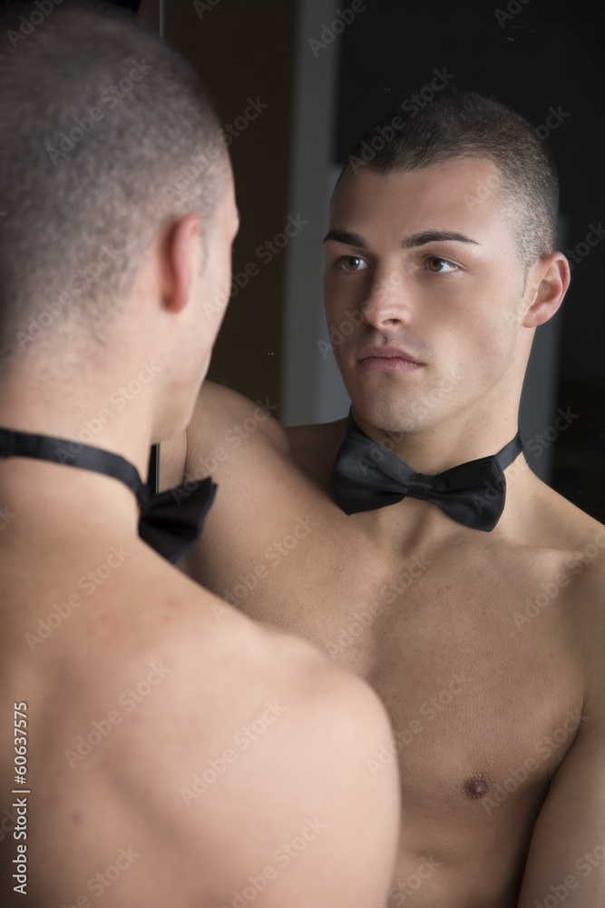 Shirtless young man with bow-tie looking at reflection in mirror Shirtless young man with bow-tie looking at reflection in mirror