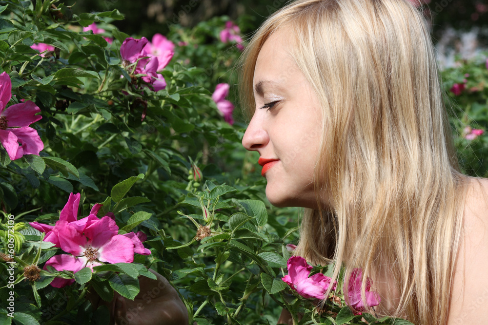 Fototapeta premium Beautiful blonde girl looks at pink flowers on bush in park