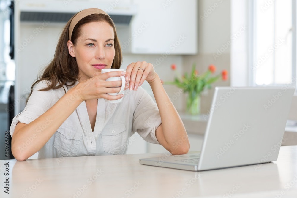 Thoughtful woman with coffee cup and laptop in kitchen Thoughtful woman with coffee cup and laptop in kitchen
