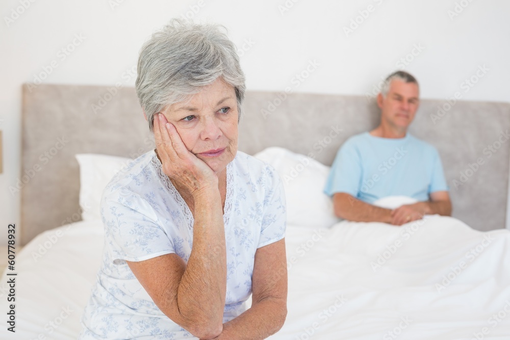 Sad senior woman on bed with husband in background Sad senior woman on bed with husband in background