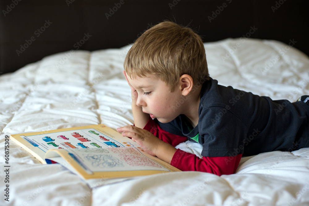 A preschool boy is studying a book while lying on a bed A preschool boy is studying a book while lying on a bed