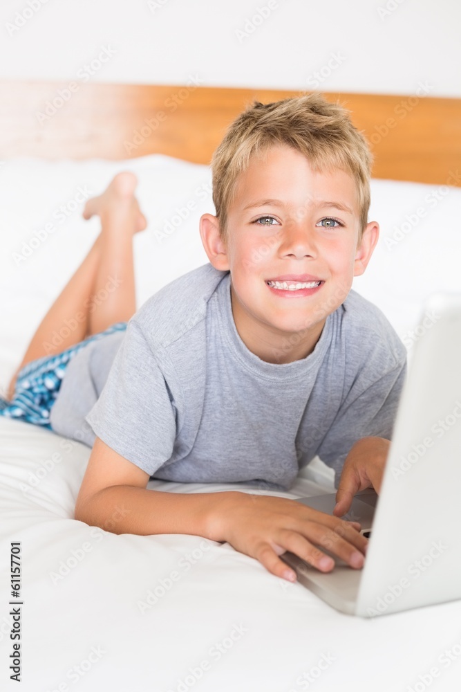 Blonde boy lying on bed using laptop smiling at camera Blonde boy lying on bed using laptop smiling at camera