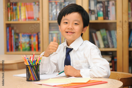Smart Asian schoolboy sitting at a desk with thumbs up