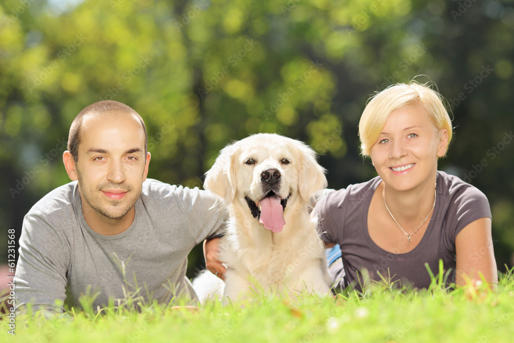 Smiling young couple lying on grass and hugging a dog in park Smiling young couple lying on grass and hugging a dog in park
