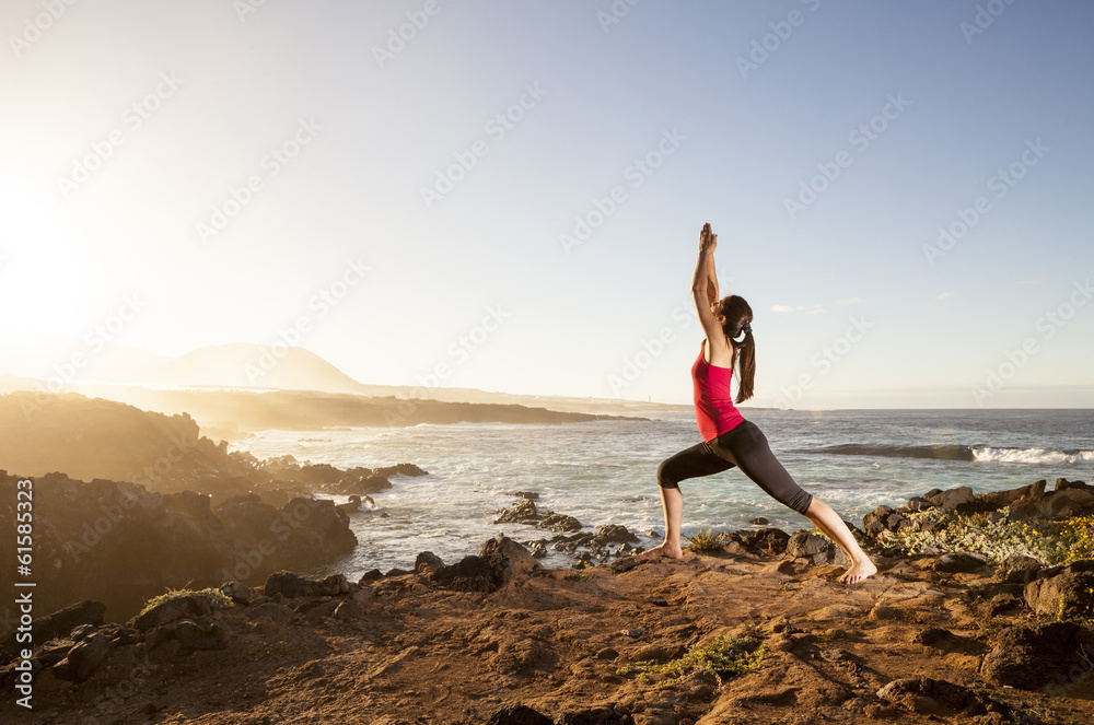 Young woman practicing yoga warrior pose near the ocean at sunse