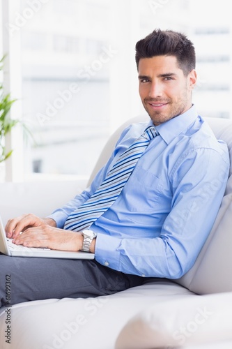 Businessman on couch using his laptop smiling at camera