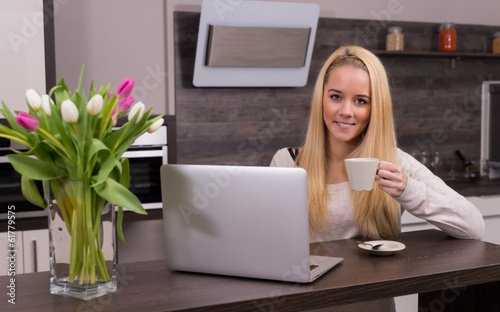 Young woman drinking coffee in kitchen with laptop