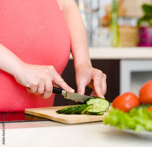 Close up pregnant woman with knife on kitchen cuts cucumber