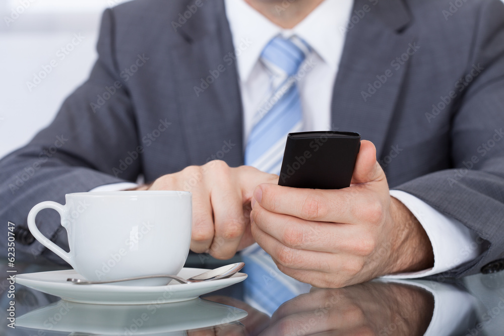 Businessperson Using Cellphone With Coffee Cup Placed At Desk Businessperson Using Cellphone With Coffee Cup Placed At Desk