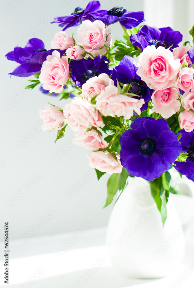 Obraz premium Bouquet of pink roses and blue anemones in white vase near the w