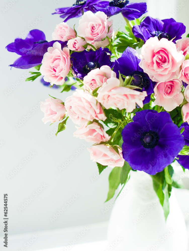 Obraz premium Bouquet of pink roses and blue anemones in white vase near the w