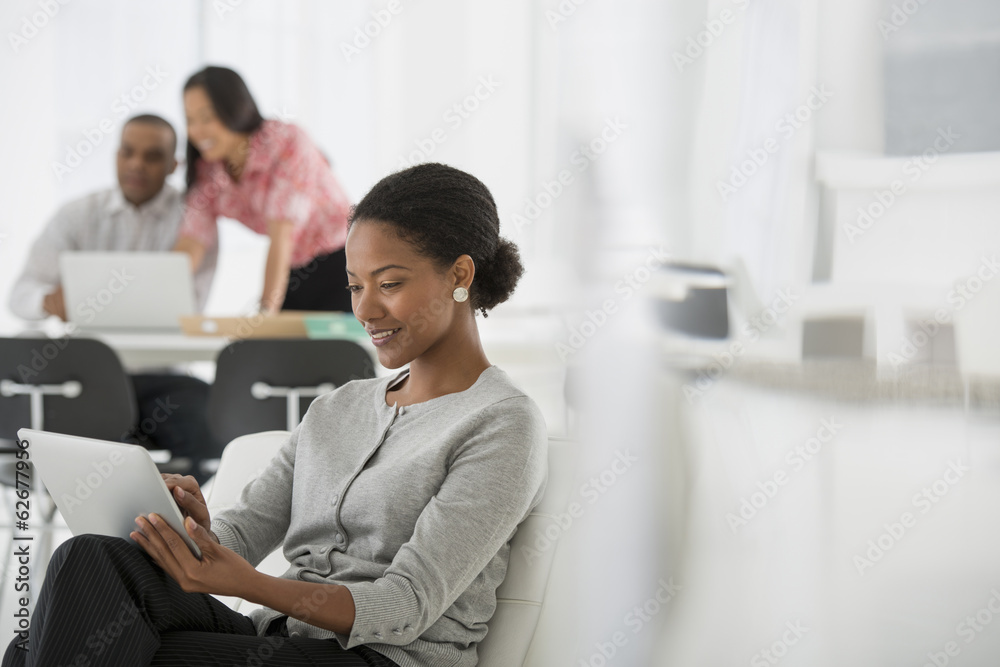 Business. A Woman Sitting Down Using A Digital Tablet. Two People In The Background Looking At A Computer Screen.