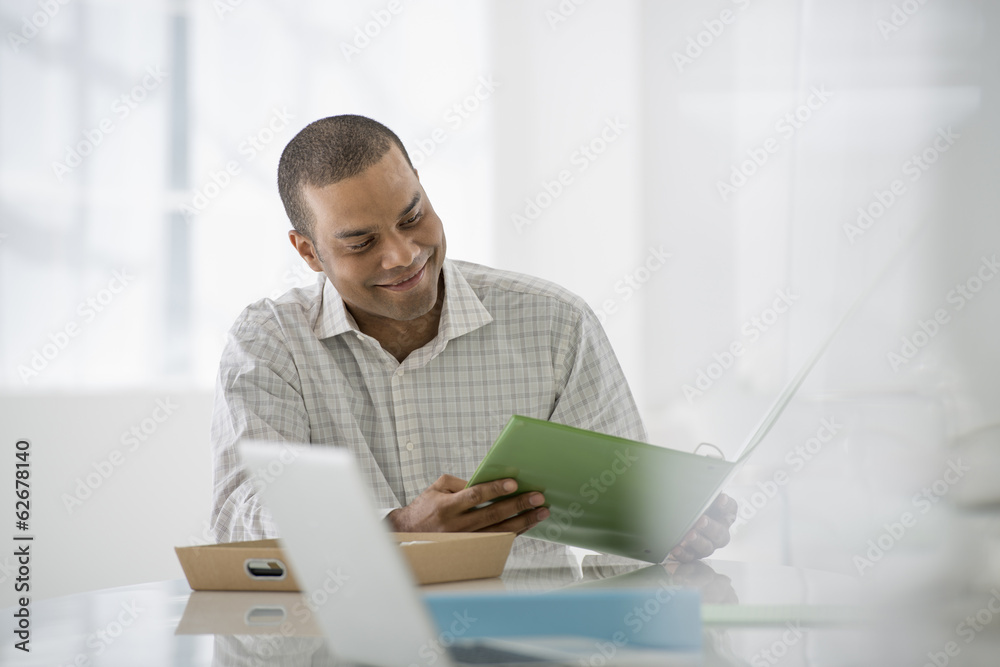 Business. A Man Seated At A Desk Opening A File Of Paperwork. A Laptop Open. Business. A Man Seated At A Desk Opening A File Of Paperwork. A Laptop Open.