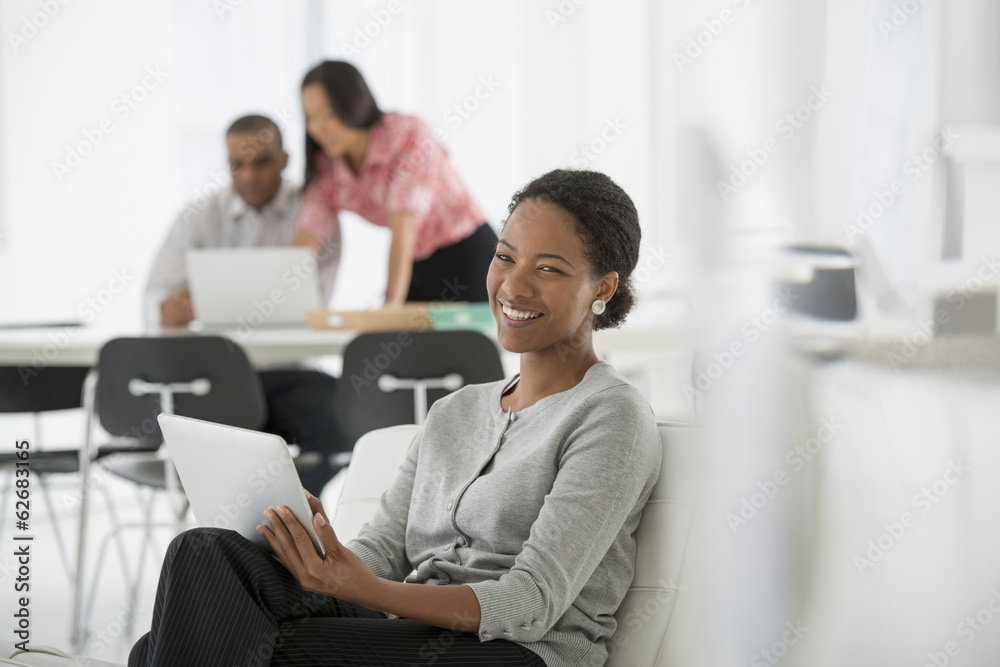Business. A Woman Sitting Down Using A Digital Tablet. Two People In The Background Looking At A Computer Screen. Business. A Woman Sitting Down Using A Digital Tablet. Two People In The Background Looking At A Computer Screen.