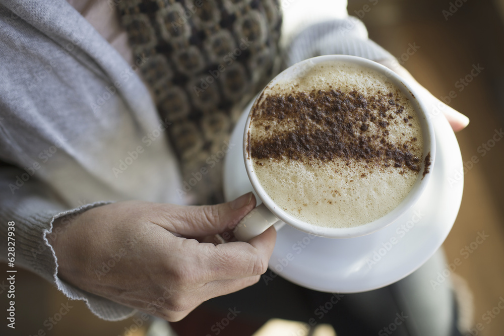 A person holding a full cup of frothy cappuccino coffee in a white china cup. Chocolate powder sprinkled in a pattern on the top. Coffee shop. A person holding a full cup of frothy cappuccino coffee in a white china cup. Chocolate powder sprinkled in a pattern on the top. Coffee shop.