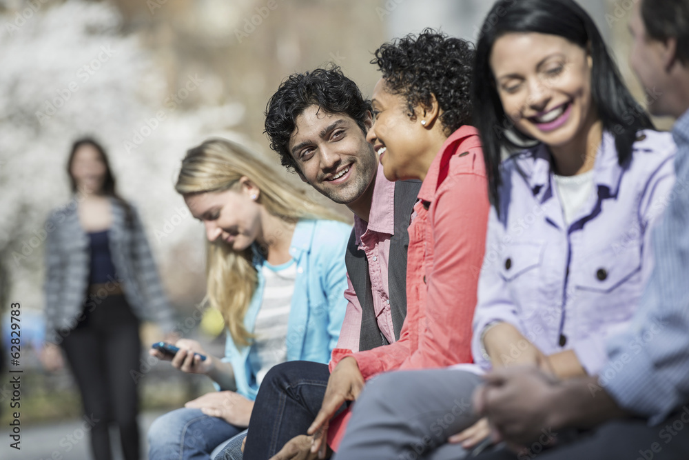 People outdoors in the city in spring time. Five people sitting in a row, one looking at a mobile phone. A woman approaching in the distance. People outdoors in the city in spring time. Five people sitting in a row, one looking at a mobile phone. A woman approaching in the distance.