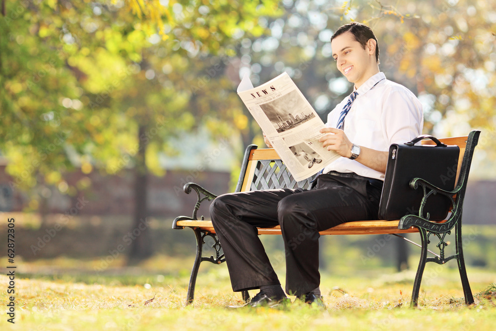 Young man seated on bench reading a newspaper on a sunny day Young man seated on bench reading a newspaper on a sunny day