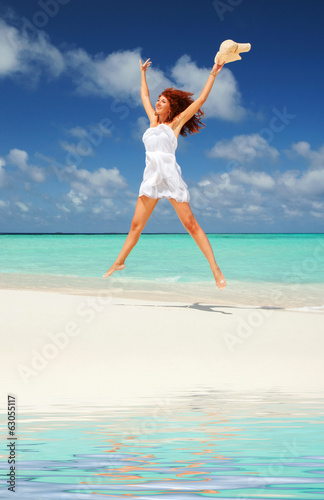 Happy young woman in white dress with hat jumping on the beach