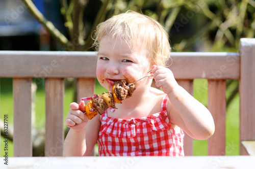 Funny toddler girl eating bbq meat outdoors in the garden