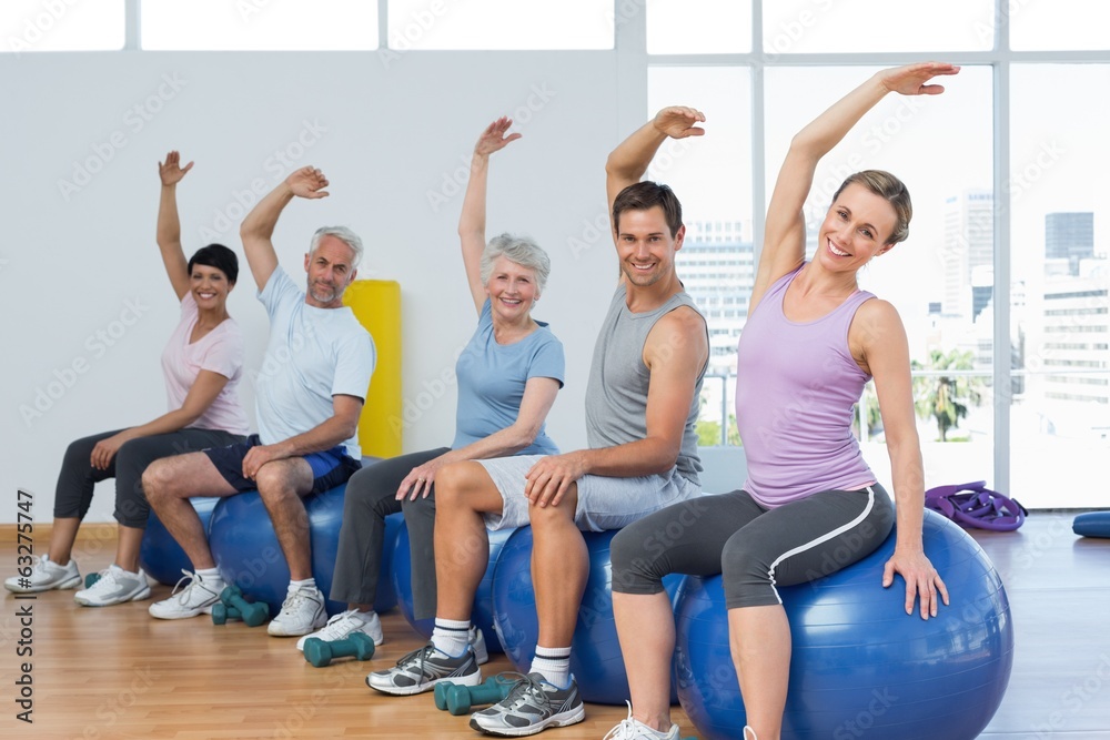 Class sitting on exercise balls and stretching hands in gym Class sitting on exercise balls and stretching hands in gym