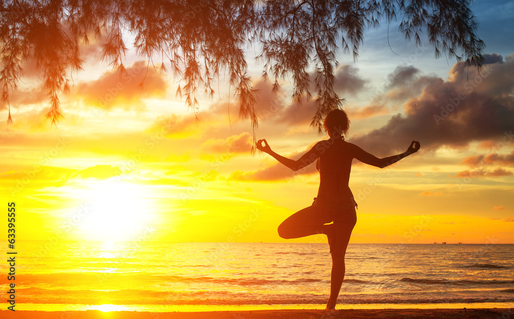 Young woman silhouette practicing yoga on beach at sunset.
