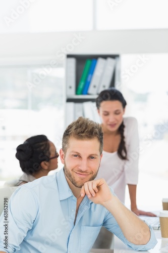 Handsome man smiling at camera while colleagues chat at desk