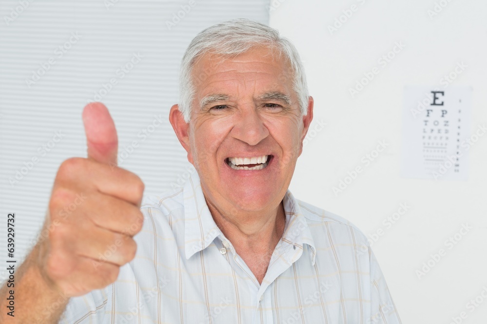 Smiling senior man gesturing thumbs up with eye chart in backgro Smiling senior man gesturing thumbs up with eye chart in backgro