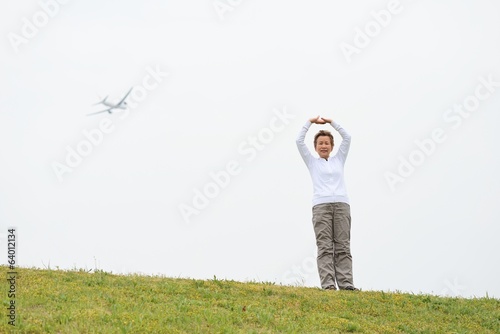白い空をバックに芝生の上でストレッチ運動をする高齢の日本人女性