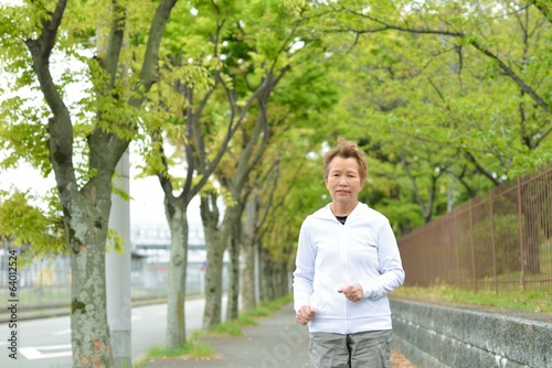 新緑の木々が植えてある歩道を健康のためにジョギングしている高齢のアジア人女性