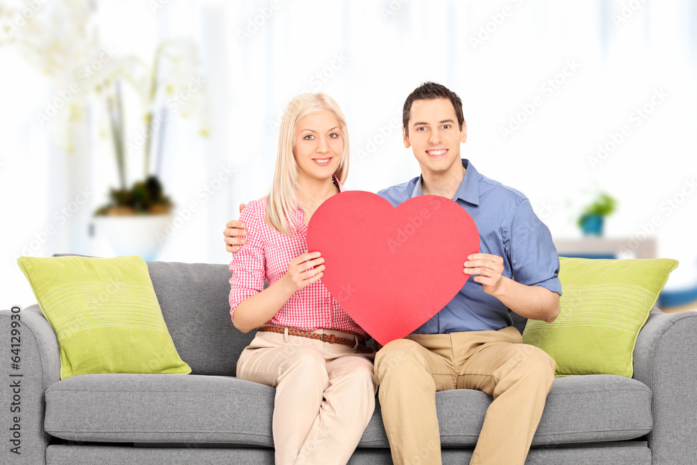 Fototapeta premium Young couple sitting at sofa and holding big red heart